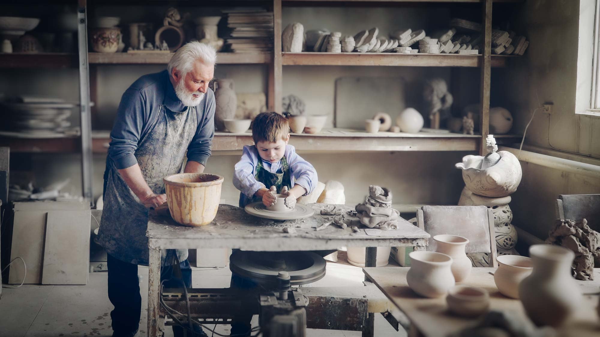 Grandfather teaching a child pottery in a workshop full of ceramic pieces — older craft studios can contain lead in glazes, pigments, and decades of accumulated dust that pose a hidden risk to visiting grandchildren