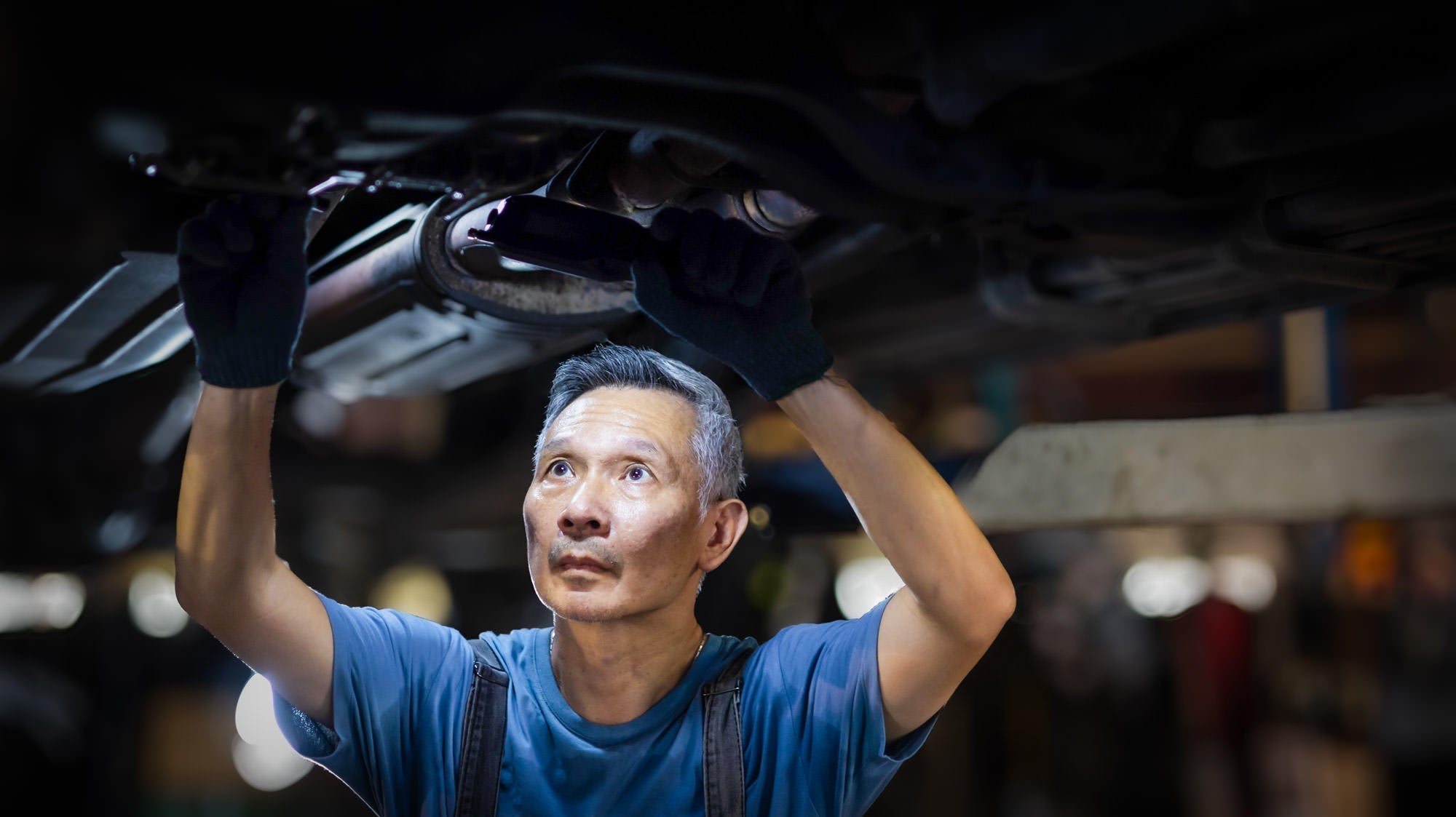 Auto mechanic working under a lifted vehicle — car repair exposes mechanics to lead in old brake dust, wheel weights, and body filler, making lead testing a workplace safety essential